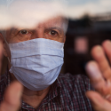 A senior man with surgical mask with hands touching window